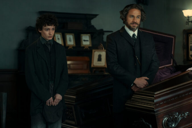 Un coup de hache dans le catalogue Netflix : Lizzie Borden est la nouvelle Monster 3 "Two men in dark, old-fashioned suits stand beside a polished wooden coffin in a dimly lit, ornate room decorated with framed photos, suggesting a 19th-century funeral or wake setting."